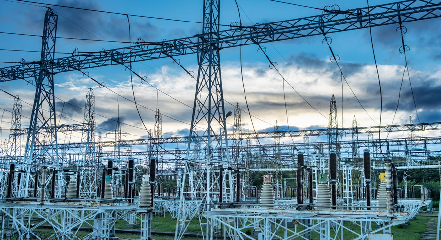An outdoor electrical substation with multiple power lines, metal towers, and transformers set against a cloudy sky at sunset.

